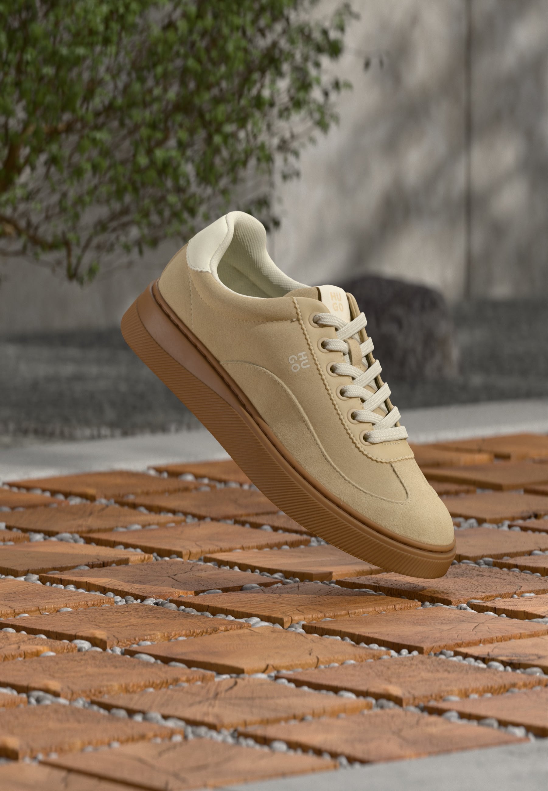 Beige Wildledersneaker mit weißen Schnürsenkeln und brauner Sohle, schwebend über einem Holzboden, im Hintergrund eine grüne Pflanze und eine graue Wand.