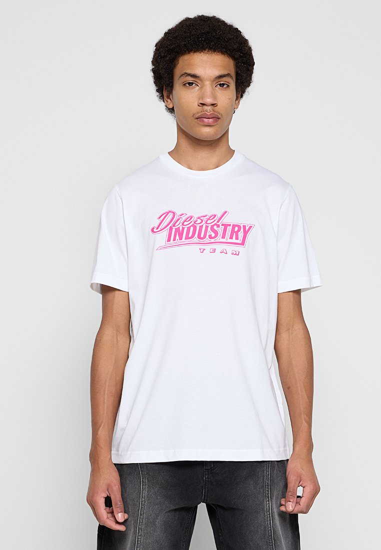 Junger Mann mit lockigem Haar, der ein weißes T-Shirt mit pinker Aufschrift "Diesel Industry Team" und ausgewaschene schwarze Jeans vor einfachem Hintergrund trägt.