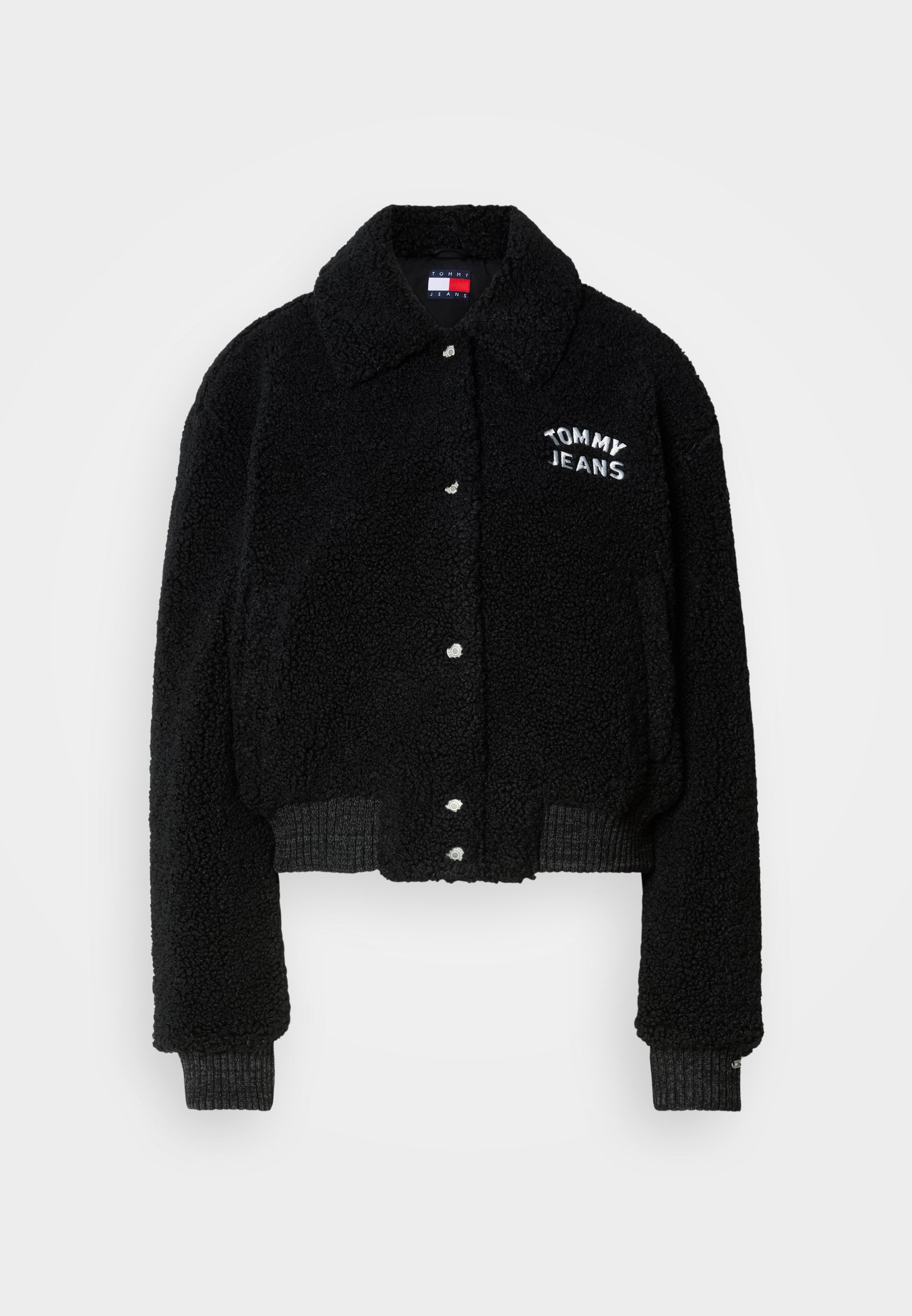 Schwarze, kurze Jacke aus flauschigem Material, mit durchgängiger Knopfleiste, Kragen und weißer "TOMMY JEANS"-Stickerei auf der Brust.