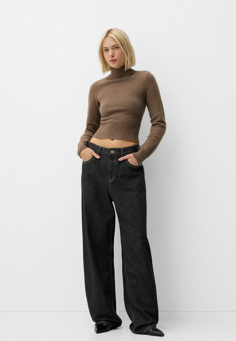 Brauner, kurzer Pullover mit geripptem Textur, kombiniert mit hochgeschnittenen schwarzen weit geschnittenen Jeans. Das Modell hat die Hände in den Taschen und trägt spitze schwarze Schuhe.