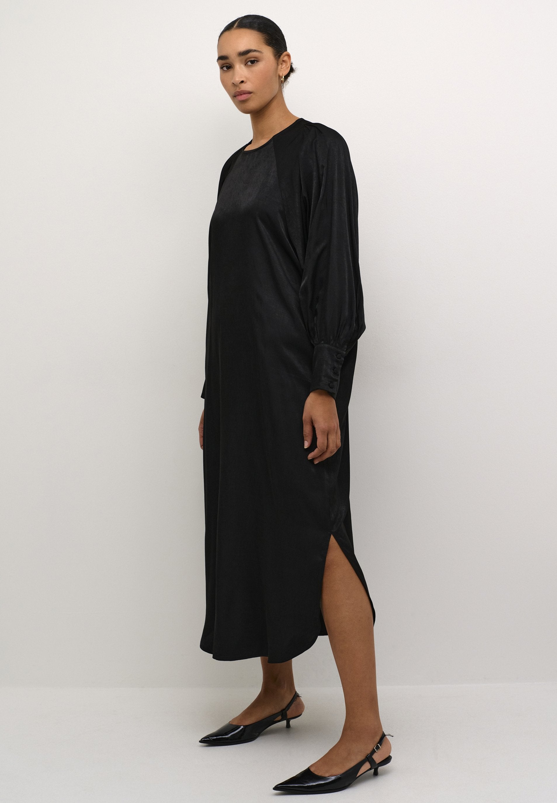 Schwarzes Satin-Kleid mit langen Ärmeln, rundem Halsausschnitt, seitlichen Schlitzen und geknöpften Manschetten. Kombiniert mit schwarzen Slingback-High Heels.