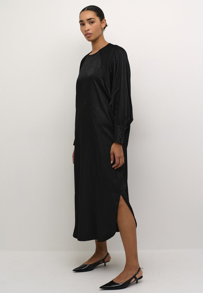 Schwarzes Satin-Kleid mit langen Ärmeln, rundem Halsausschnitt, seitlichen Schlitzen und geknöpften Manschetten. Kombiniert mit schwarzen Slingback-High Heels.