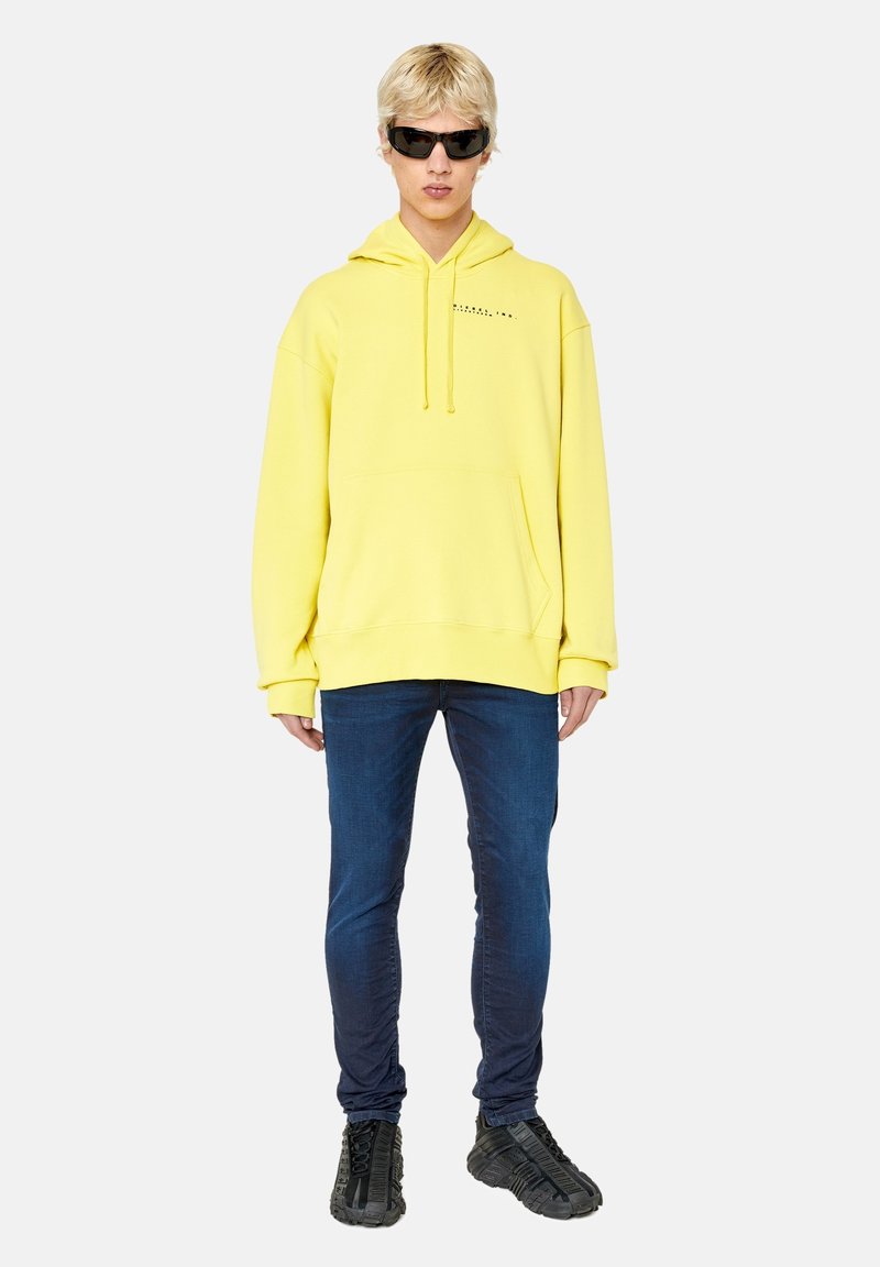 Gelbes Kapuzensweatshirt mit Fronttasche, kombiniert mit dunklen Slim-Fit-Jeans und schwarzen Sportschuhen. Das Modell trägt Sonnenbrillen.