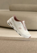 Weißer und mauvefarbener Sportschuh mit Luftpolstersohle, Skechers-Logo an der Seite, dargestellt auf einer strukturierten Oberfläche mit beigem Sofa im Hintergrund.
