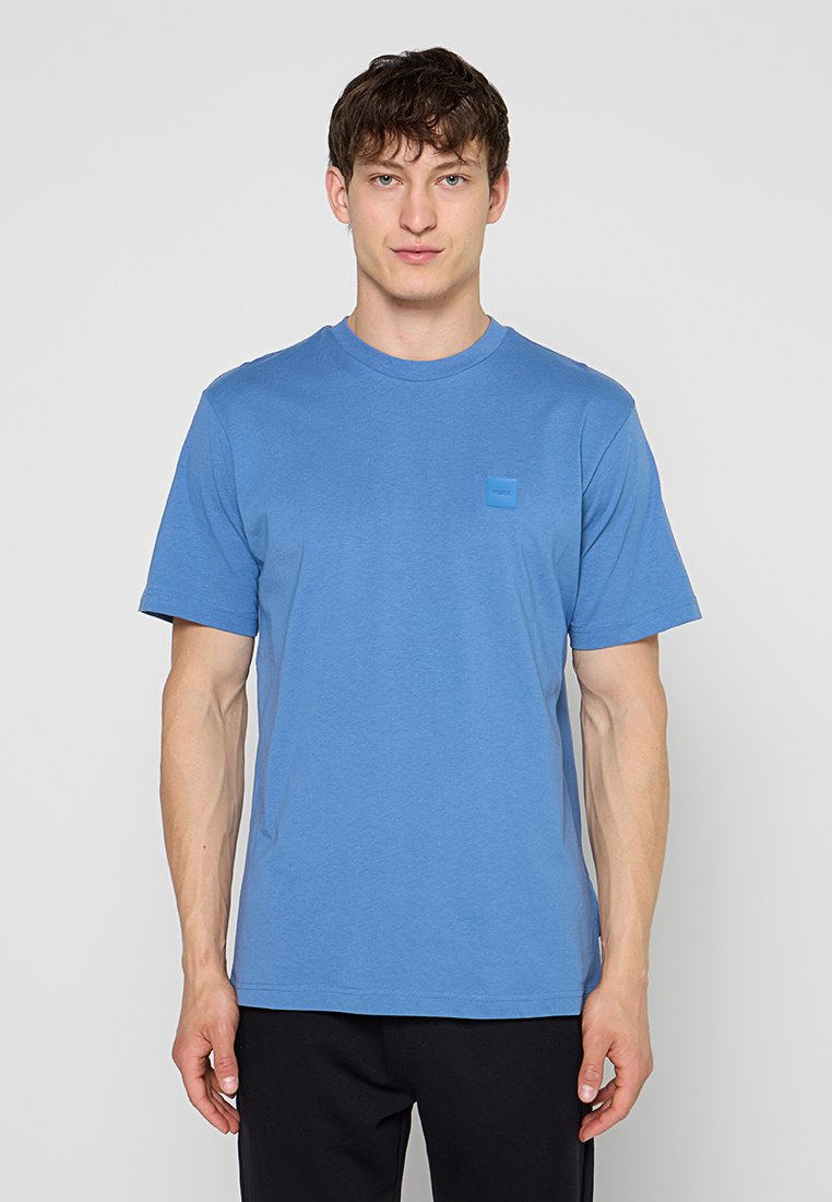 Junger Mann, der frontal steht und ein schlichtes blaues T-Shirt mit Rundhalsausschnitt und schwarze Hose vor einem hellgrauen Hintergrund trägt.