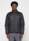 Mann trägt eine schwarze, gesteppte Jacke mit Reißverschluss und hohem Kragen sowie graue Hose, steht vor einem schlichten hellen Hintergrund.