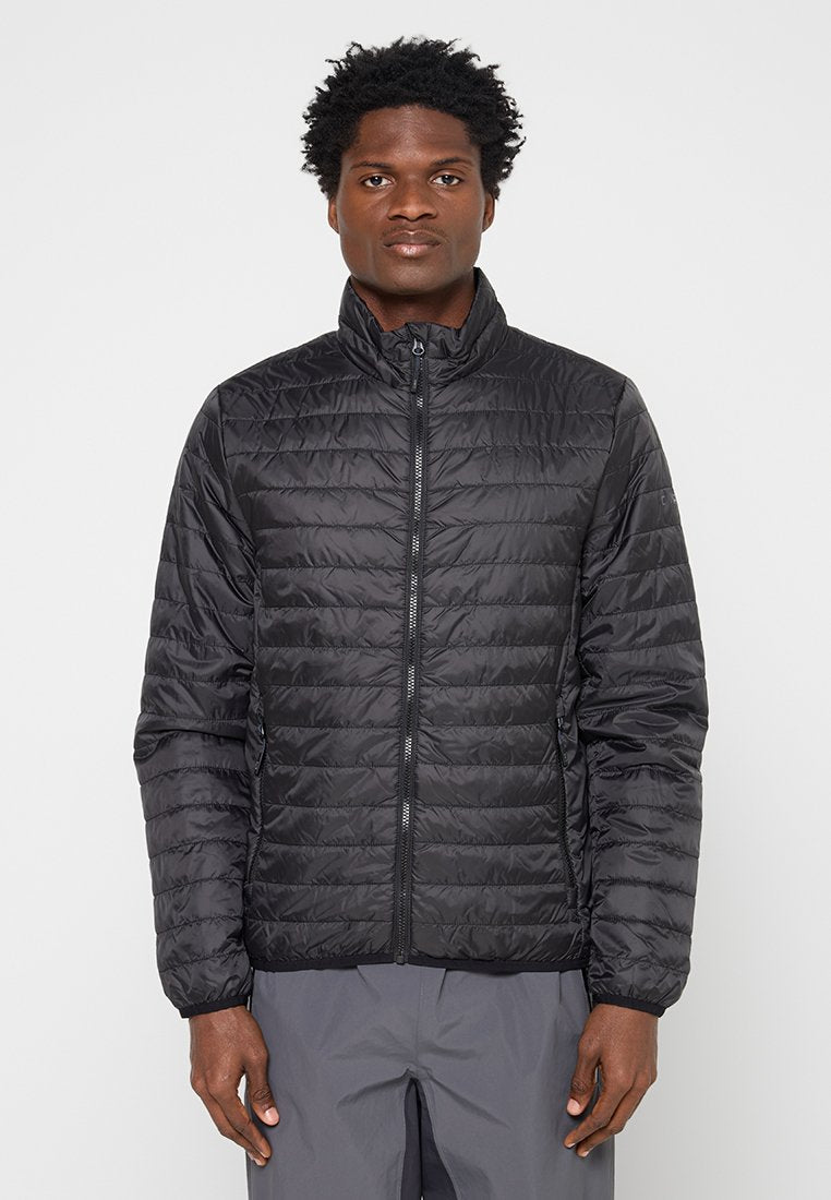 Mann trägt eine schwarze, gesteppte Jacke mit Reißverschluss und hohem Kragen sowie graue Hose, steht vor einem schlichten hellen Hintergrund.