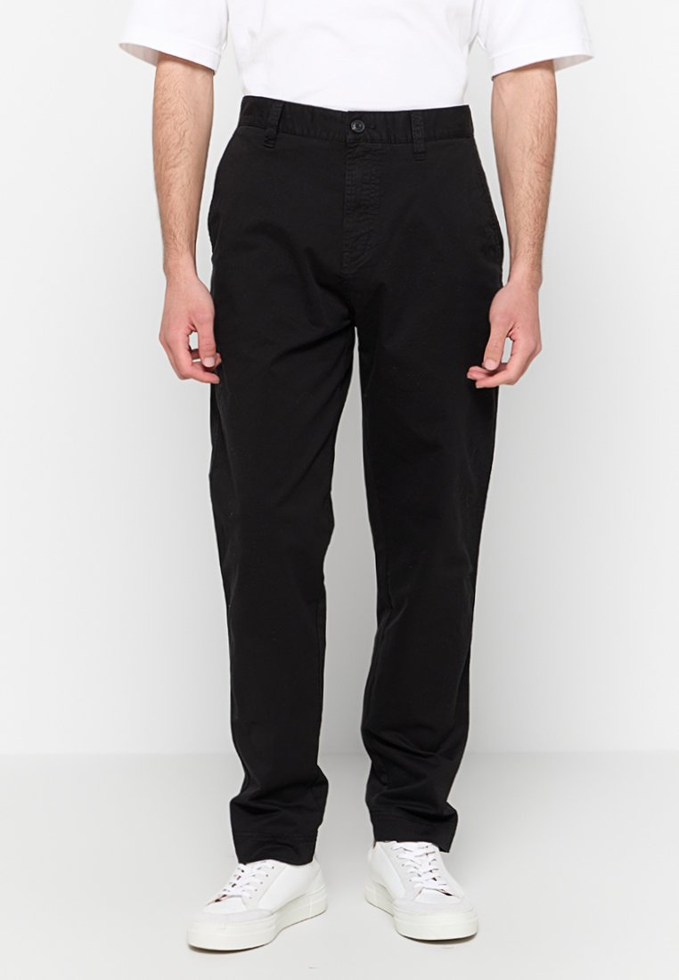 Schwarze gerade geschnittene Hosen mit einem Frontknopfverschluss, Gürtelschlaufen und Seitentaschen, aus glattem Baumwollstoff. Kombiniert mit weißen Sneakern.