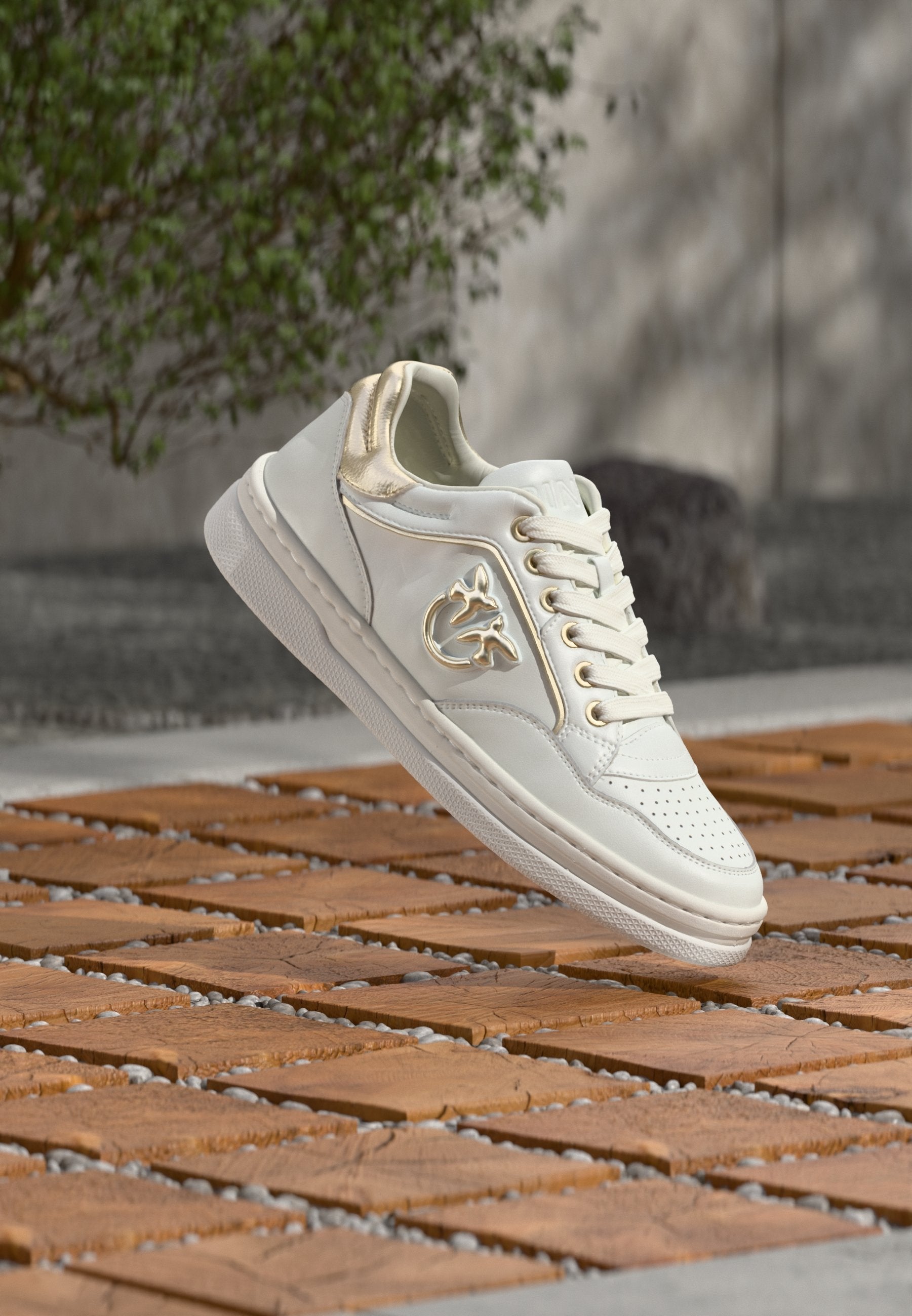 Weißer Sneaker mit goldenen Akzenten und Schnürdesign, der über Holzfliesen im Freien nahe Grünpflanzen und einer Betonmauer schwebt.