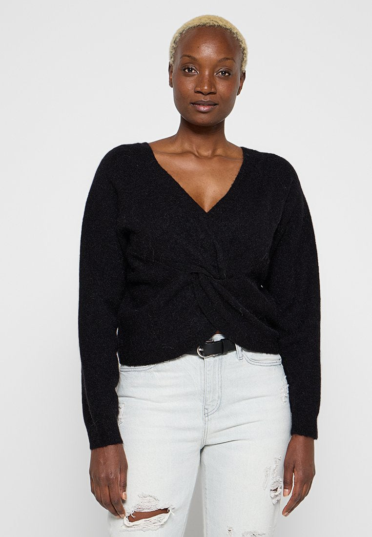 Schwarzer Strickpullover mit tiefem V-Ausschnitt und gedrehtem Vorderteil. Kombiniert mit hellblauen, ausgewaschenen Jeans und einem schwarzen Gürtel.
