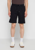 Mann trägt schwarze Shorts mit Kordelzug, weiße Socken und weiße Sneakers, steht vor einem einfarbigen hellen Hintergrund.