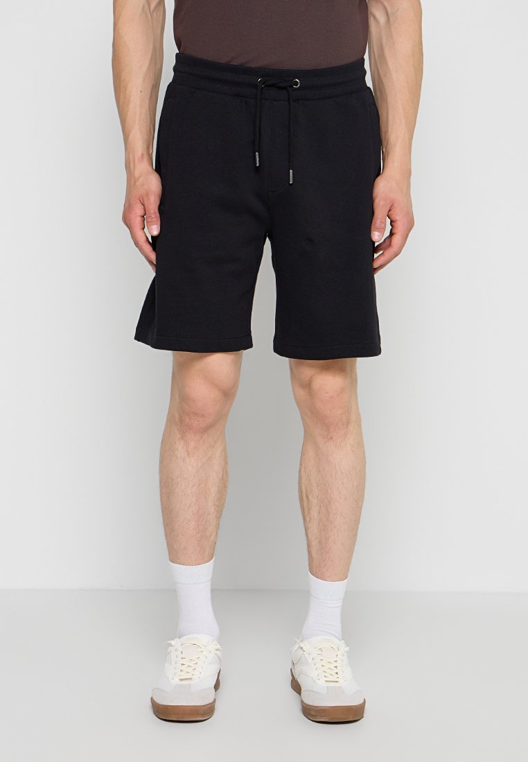 Mann trägt schwarze Shorts mit Kordelzug, weiße Socken und weiße Sneakers, steht vor einem einfarbigen hellen Hintergrund.