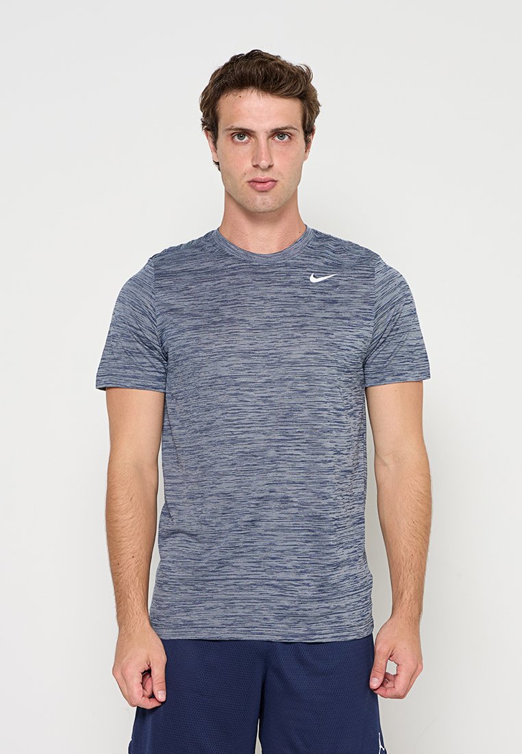 Junger Mann, der nach vorne schaut, trägt ein blaues meliertes Nike-T-Shirt und dunkelblaue Shorts vor einem einfarbigen hellen Hintergrund.