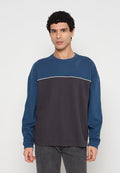 Mann mit lockigem Haar, der einen blau-dunkelgrauen langärmligen Sweatshirt mit Farbblockmuster und dunkelgraue Hosen trägt, vor einem einfarbigen Hintergrund steht.