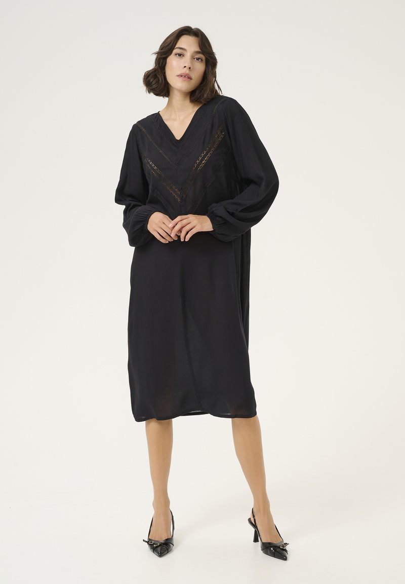 Schwarzes knielanges Kleid mit langen Ärmeln und V-Ausschnitt, das mit Spitze im Zickzackmuster entlang der Brust und Ärmel verziert ist.