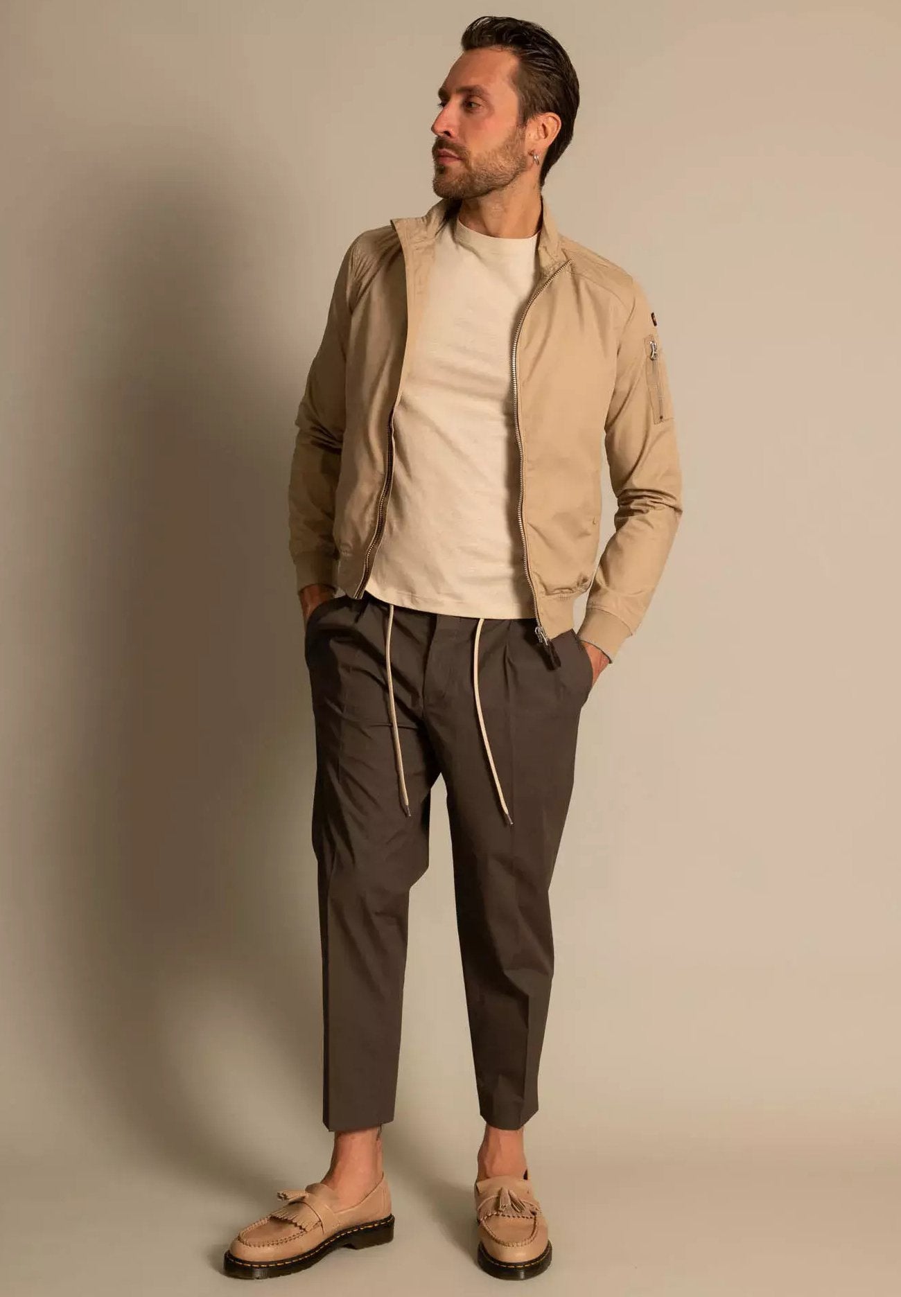 Beige Bomberjacke, cremiges Hemd, dunkelgraue Hose mit Kordelzug und hellbraune Slipper mit dekorativen Akzenten. Minimalistisches Design.