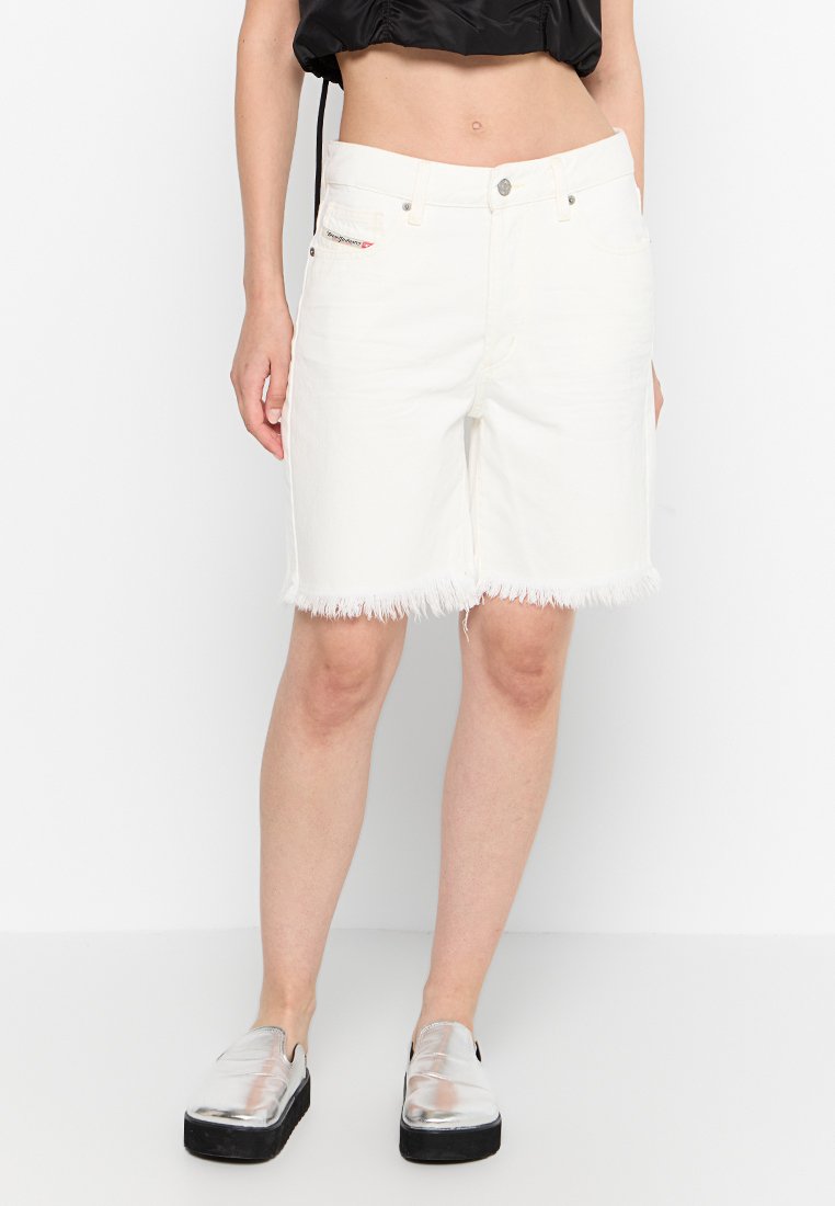 Frau in weißen, ausgefransten Denim-Shorts, schwarzem Cropped-Top und silbernen Slippern, die vor einem schlichten weißen Hintergrund steht.