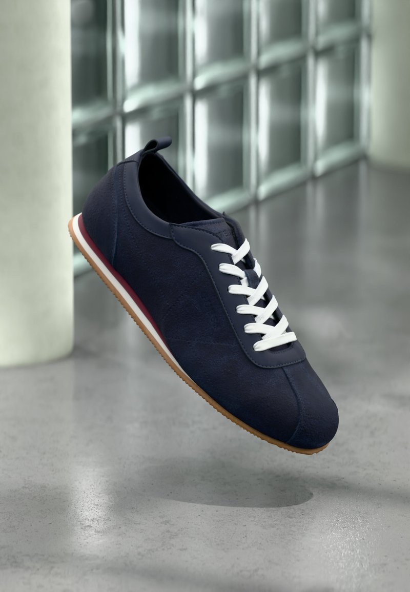 Marineblaue Freizeitsneaker mit weißen Schnürsenkeln, Gummisohle und rotem Streifen, schwebend über einem glänzenden Betonboden in der Nähe von Glasbausteinen.