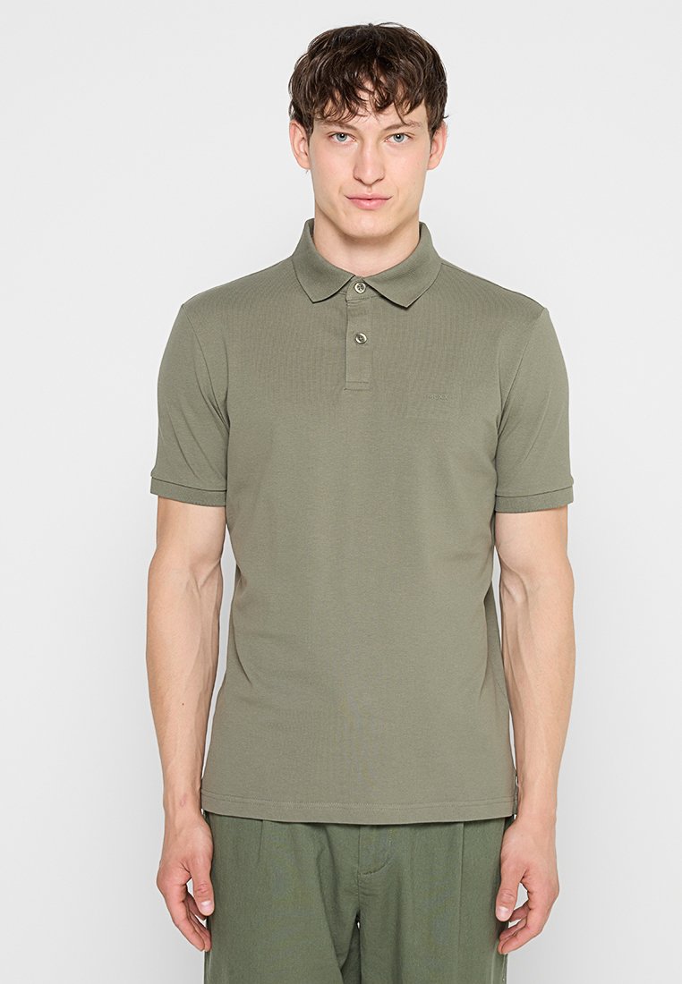 Mann mit kurzen braunen Haaren, der ein olivgrünes kurzärmeliges Poloshirt und passende Hose vor einem schlichten weißen Hintergrund trägt.