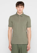 Mann mit kurzen braunen Haaren, der ein olivgrünes kurzärmeliges Poloshirt und passende Hose vor einem schlichten weißen Hintergrund trägt.