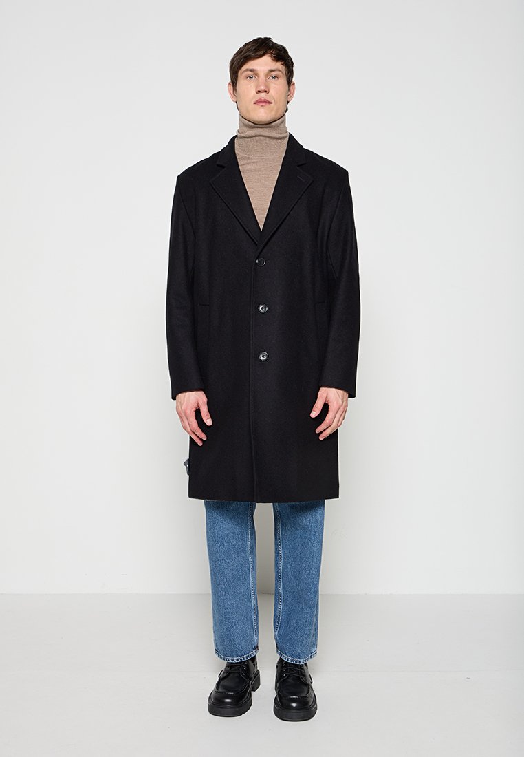Ein Mann trägt einen schwarzen, geknöpften Mantel über einem beigefarbenen Rollkragenpullover, blaue Jeans und schwarze Schuhe und steht vor einem schlichten weißen Hintergrund.