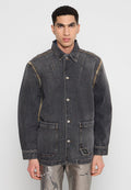 Junger Mann mit kurzen, lockigen Haaren, der eine dunkelgraue, knöpfbare Jeansjacke und metallisch gemusterte Hosen trägt, steht vor einem schlichten Hintergrund.