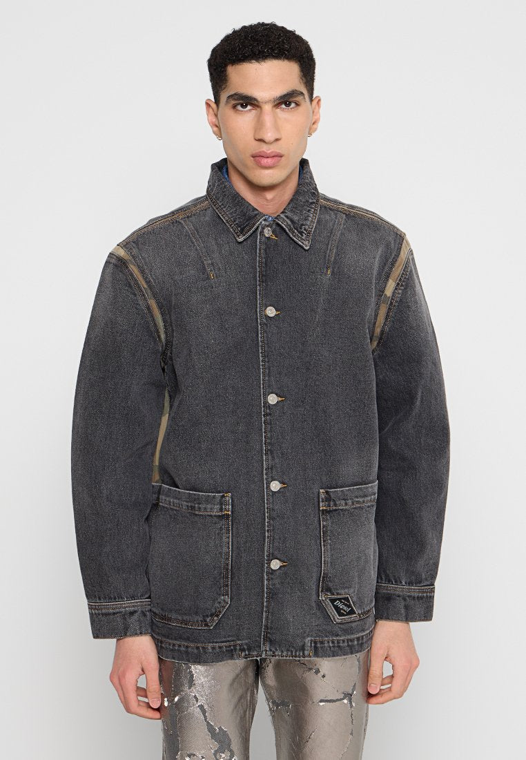 Junger Mann mit kurzen, lockigen Haaren, der eine dunkelgraue, knöpfbare Jeansjacke und metallisch gemusterte Hosen trägt, steht vor einem schlichten Hintergrund.