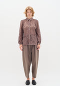 Braune Spitzenbluse mit Kragen und langen Ärmeln, die komplizierte Blumenmuster aufweist. Kombiniert mit lockeren, grauen Hosen und schwarzen Slippern.