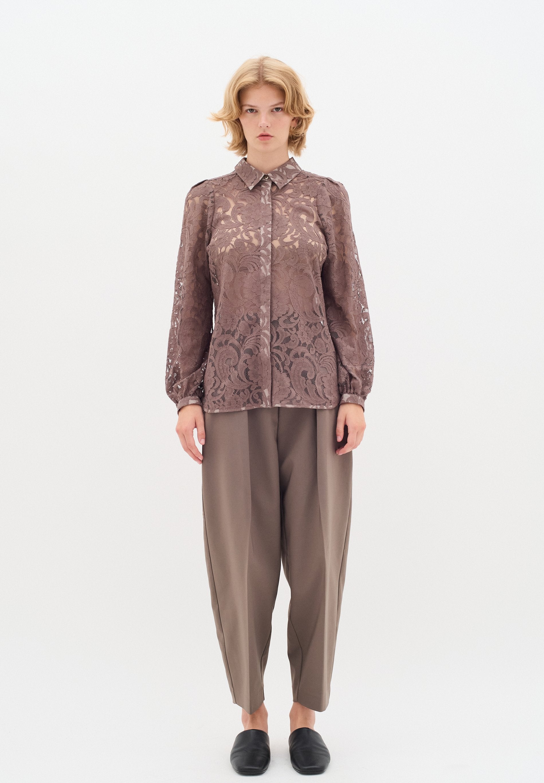 Braune Spitzenbluse mit Kragen und langen Ärmeln, die komplizierte Blumenmuster aufweist. Kombiniert mit lockeren, grauen Hosen und schwarzen Slippern.