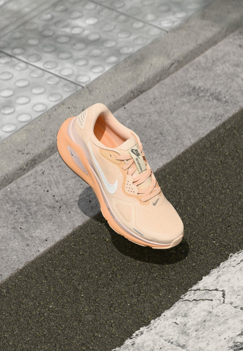 Helle pfirsichfarbene Nike-Laufschuhe mit weißem Swoosh auf grauem Asphalt neben dem Bordstein und taktilen Pflastersteinen.