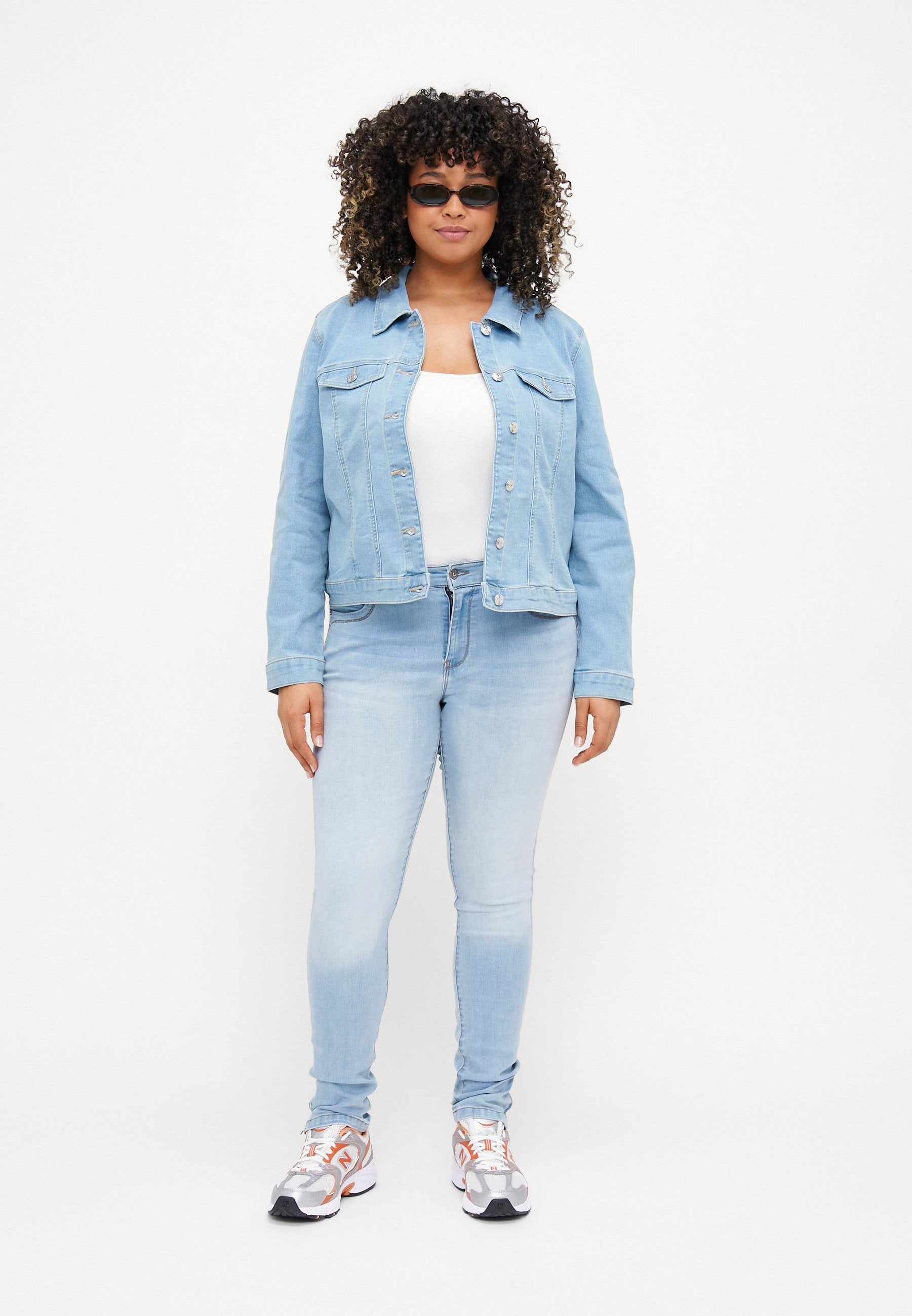Helle blaue Jeansjacke mit zwei Fronttaschen, Knopfverschluss und darunter einem weißen Oberteil. Helle blaue Skinny-Jeans und sportliche Sneakers.
