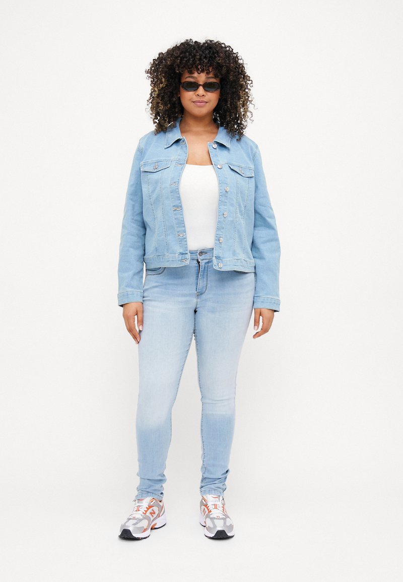 Helle blaue Jeansjacke mit zwei Fronttaschen, Knopfverschluss und darunter einem weißen Oberteil. Helle blaue Skinny-Jeans und sportliche Sneakers.
