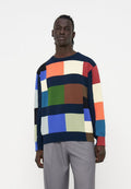 Junger Mann trägt einen bunten Patchwork-Pullover mit blauen, grünen, orangen, roten und weißen Blöcken, kombiniert mit grauen Hosen, steht vor weißem Hintergrund.