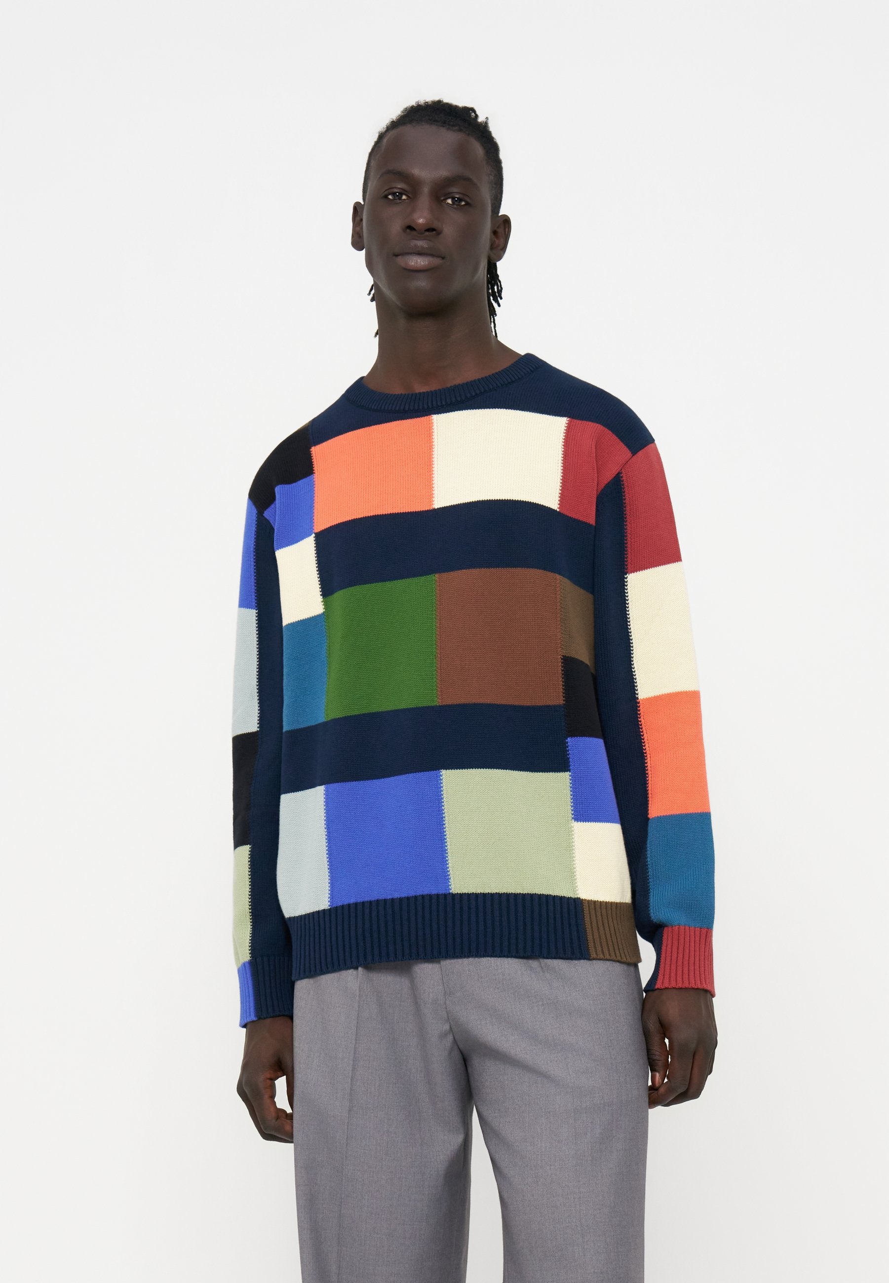 Junger Mann trägt einen bunten Patchwork-Pullover mit blauen, grünen, orangen, roten und weißen Blöcken, kombiniert mit grauen Hosen, steht vor weißem Hintergrund.