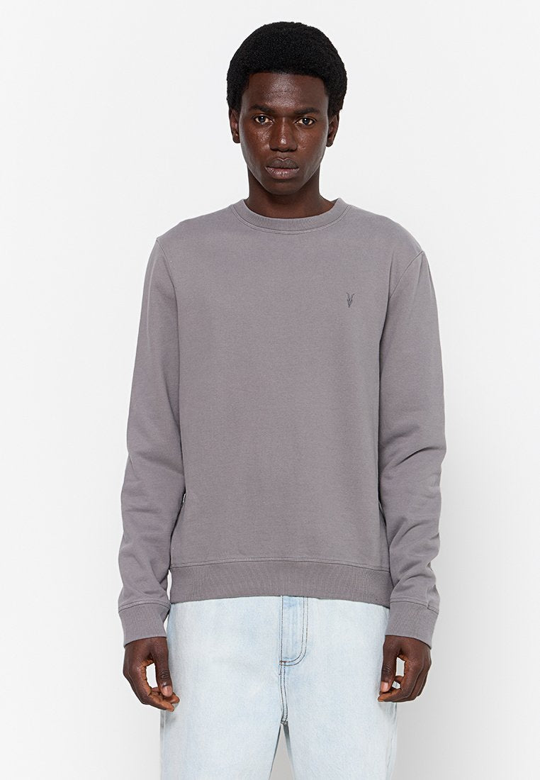 Mann mit kurzem lockigem Haar, der einen einfachen grauen Langarmpullover und hellblaue Jeans trägt, vor einem weißen Hintergrund steht.