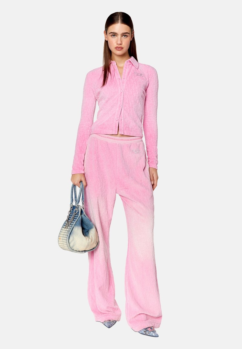 Eine Frau trägt eine rosa samtige, geknöpfte Jacke und passende weit geschnittene Hosen, hält eine blau-weiße Handtasche und steht vor einem weißen Hintergrund.