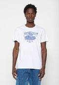 Mann mit kurzen lockigen Haaren, der ein weißes T-Shirt mit blauem Motorrad-Grafik und Text trägt, kombiniert mit blauen Denim-Jeans.