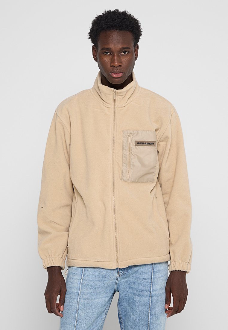 Beige Zip-Jacke aus weichem Fleece-Material mit einer vorderen Tasche mit Reißverschluss und Logodetails. Elastische Bündchen und Saum.