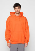 Mann trägt hellorange Kapuzenpullover mit Fronttasche und kleinem weißem Logo, steht vor hellem einfarbigem Hintergrund.