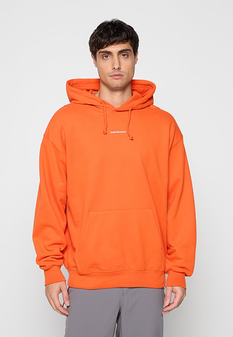 Mann trägt hellorange Kapuzenpullover mit Fronttasche und kleinem weißem Logo, steht vor hellem einfarbigem Hintergrund.