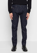 Mann trägt dunkelblaue, gerade geschnittene Jeans, einen schwarzen Pullover und schwarze Lederstiefel, steht vor einem schlichten weißen Hintergrund.