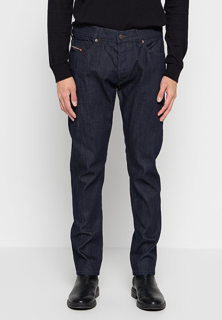 Mann trägt dunkelblaue, gerade geschnittene Jeans, einen schwarzen Pullover und schwarze Lederstiefel, steht vor einem schlichten weißen Hintergrund.