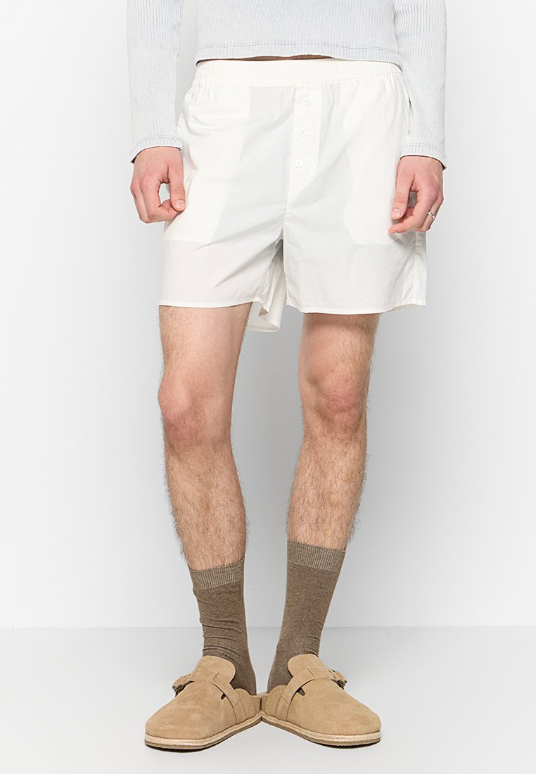 Weiße Baumwollboxershorts mit Knopfverschluss, elastischem Bund und seitlichen Schlitzen. Getragen mit beigen Slippers und braunen Socken.