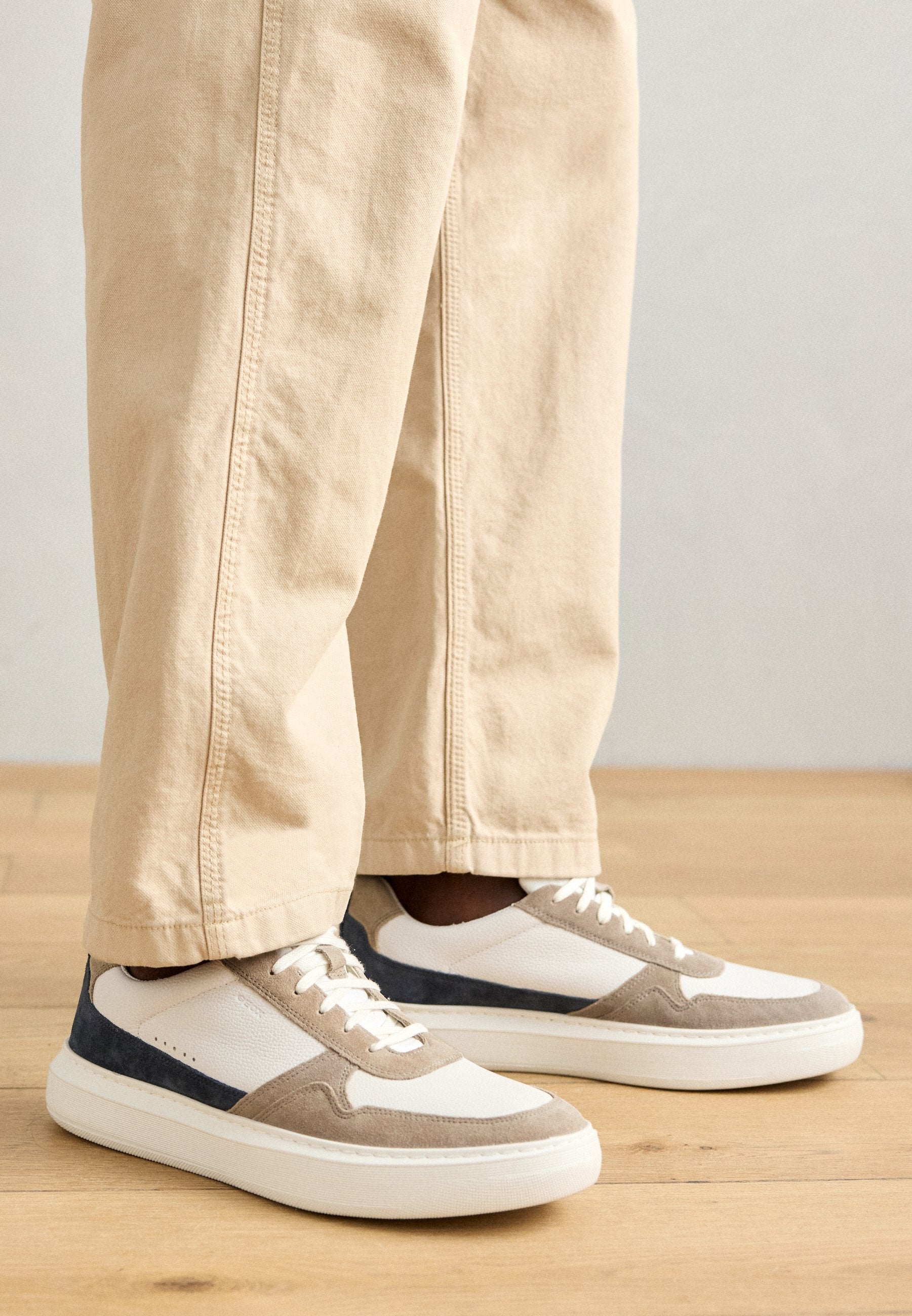 Sneaker mit einem Obermaterial aus weißem Leder, beigen Wildledereinsätzen und marineblauen Details. Dicke weiße Sohle und Stoffsenkel. Kombiniert mit beigen Hosen.