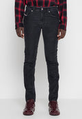 Mann trägt eng anliegende schwarze Cordjeans, ein rot-schwarz kariertes Hemd und glänzende dunkelrote Turnschuhe, steht vor einem schlichten Hintergrund.