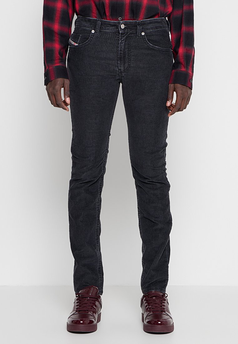 Mann trägt eng anliegende schwarze Cordjeans, ein rot-schwarz kariertes Hemd und glänzende dunkelrote Turnschuhe, steht vor einem schlichten Hintergrund.