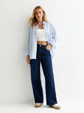 Hellblau-weiß gestreiftes Kurzarm-Hemd, getragen über einem weißen Cropped-Top, kombiniert mit hochtaillierten, dunkelblauen weit geschnittenen Jeans und beigen Schuhen.
