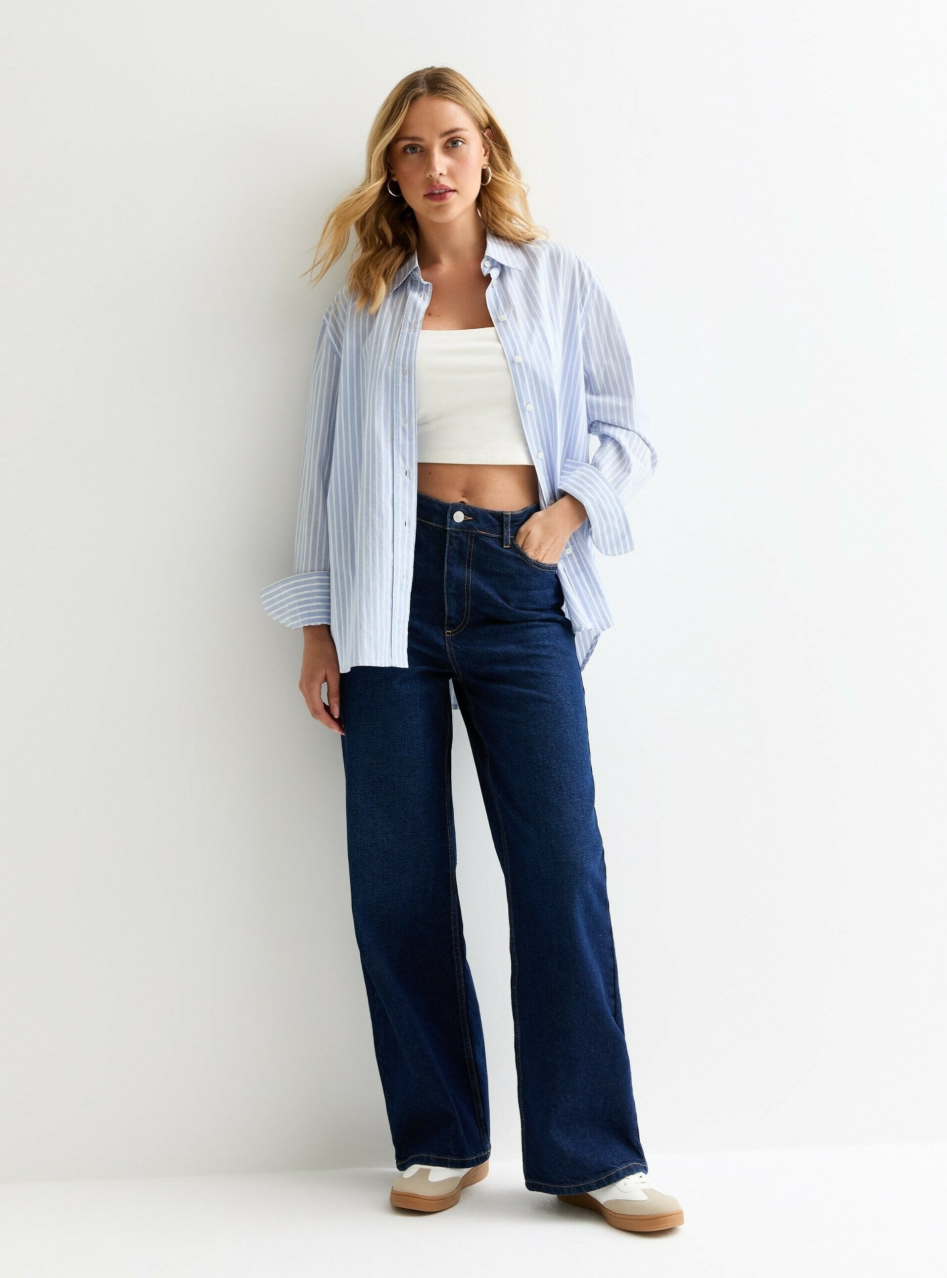 Hellblau-weiß gestreiftes Kurzarm-Hemd, getragen über einem weißen Cropped-Top, kombiniert mit hochtaillierten, dunkelblauen weit geschnittenen Jeans und beigen Schuhen.