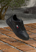 Schwarze Low-Top-Sneaker mit Tommy Jeans Logo, schwarzen Schnürsenkeln, über Holzfliesen mit grauen Kieseln und verschwommenem Baumhintergrund ausgestellt.