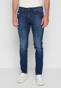 Mann in schmal geschnittenen dunkelblauen Jeans mit dezenten Auswaschungen und weißen Sneakers, vor einem einfarbigen hellen Hintergrund stehend.